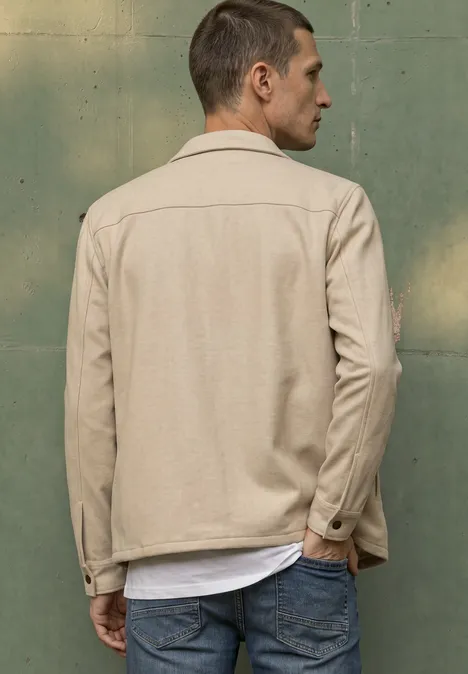 Ein Mann von hinten mit beiger Jacke und blauer Jeans vor grünem Hintergrund.