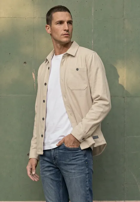 Mann in beigefarbener Overshirt, weißem T-Shirt und Jeans vor grünem Hintergrund.