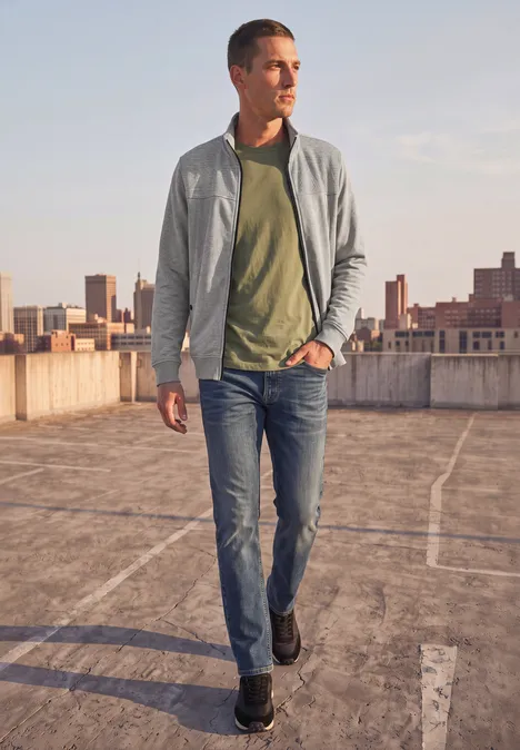 Een man in jeans, groen T-shirt en grijze jas loopt over een parkeerdak met stad op de achtergrond.