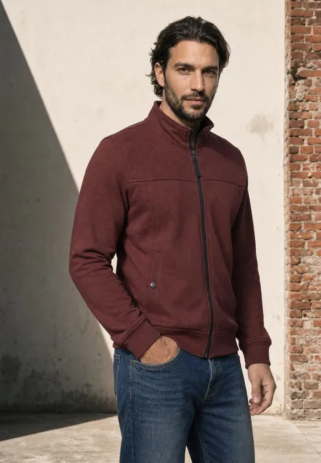 Ein Mann mit dunklem Haar und Bart steht in Jeans und bordeauxrotem Fleecejacke vor einer Wand.
