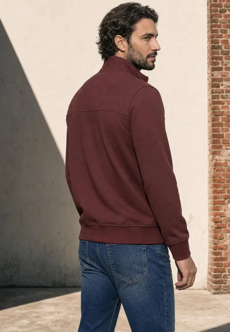 Ein Mann in weinrotem Pullover und Jeans steht vor einer Wand und blickt zur Seite.