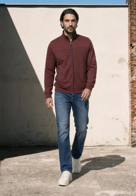 Ein Mann mit dunkelroter Jacke, Jeans und weißen Sneakern geht vor einer Wand mit Schatten.