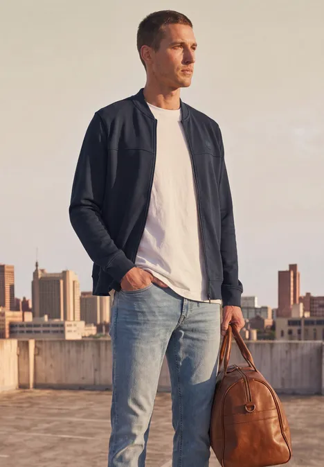 Blouson en piqué double face à fermeture zip dark ink blue Ein Mann in weißem T-Shirt, schwarzer Bomberjacke und Jeans hält eine braune Ledertasche auf einer Dachterrasse.