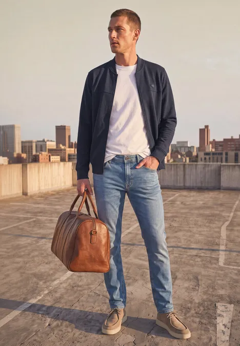 Blouson en piqué double face à fermeture zip dark ink blue Ein Mann in Jeans, weißem T-Shirt und leichter Jacke mit Lederreisetasche auf einem Parkdeck vor der Stadt.