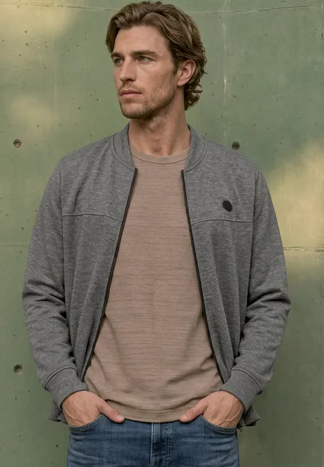 Doubleface Piqué Blouson mit Zipper metal grey melange Ein junger Mann in grauer Bomberjacke und beige T-Shirt lehnt mit den Händen in den Taschen an einer grünen Wand.