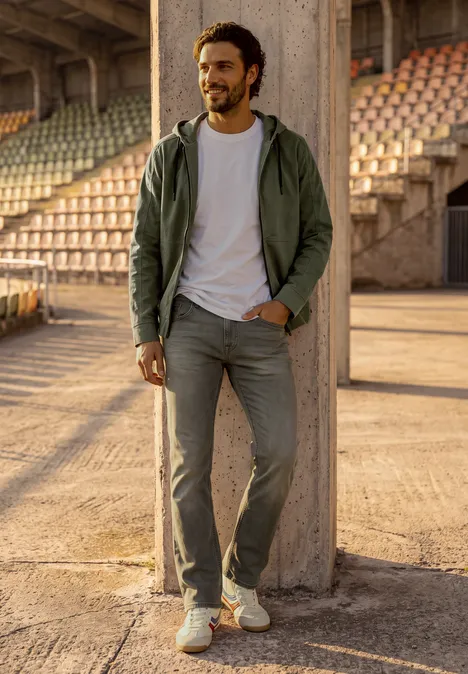 Glimlachende man in jeans en jas leunend tegen een betonnen pilaar in een stadion.