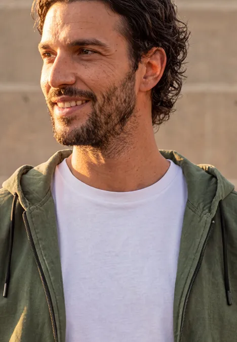 Een glimlachende man met baard draagt een wit T-shirt en een groen capuchonvest buiten.