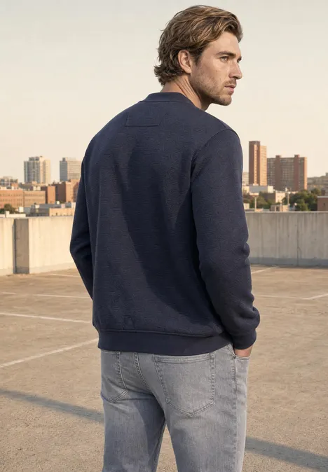 Un homme de dos sur un toit-terrasse urbain, vêtu d’un pull bleu et d’un jean, avec la ville en arrière-plan.