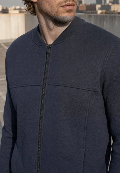 Un homme portant un pull zippé bleu foncé sur un toit avec une ville en arrière-plan.