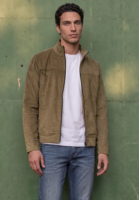 Un jeune homme porte une veste en velours côtelé marron, un t-shirt blanc et un jean, devant un mur vert.