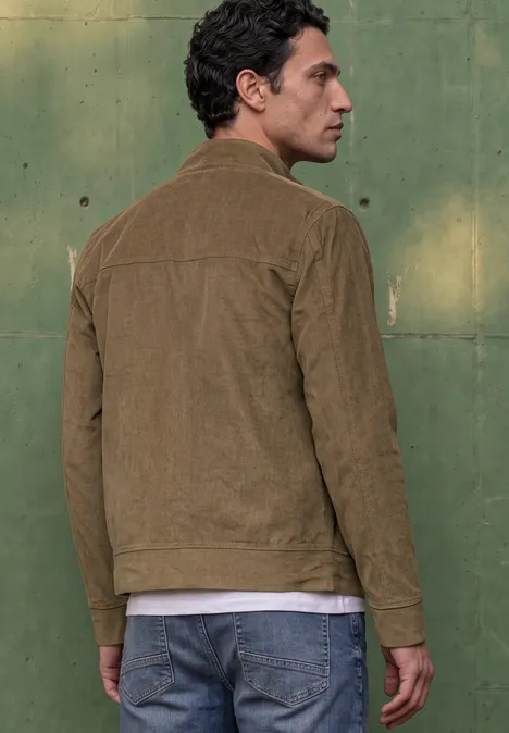 Un homme devant un mur vert, portant une veste en velours marron et un jean bleu, regarde par-dessus son épaule.