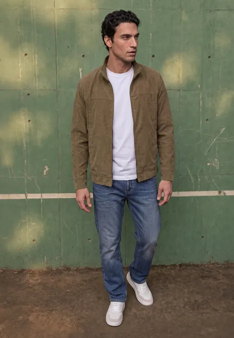 Un homme en veste beige, t-shirt blanc, jeans bleus et baskets blanches se tient devant un mur vert.