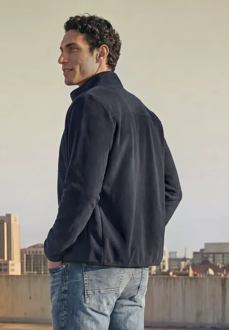Ein Mann steht auf einem Dach mit städtischer Kulisse, trägt eine dunkle Fleecejacke und Jeans, schaut seitlich.