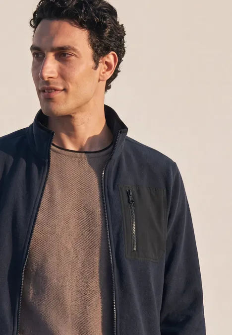 Ein Mann mit dunkler Jacke und grauem T-Shirt blickt seitlich vor neutralem Hintergrund.