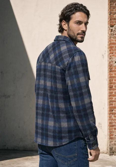 Ein bärtiger Mann im blauen karierten Hemd steht seitlich vor einer Wand und blickt weg.
