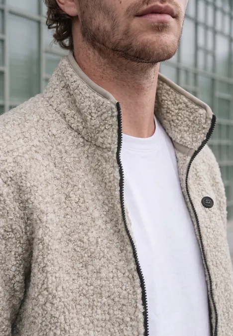 Ein Mann trägt ein weißes T-Shirt und eine beige Fleecejacke vor einer Glasfront.