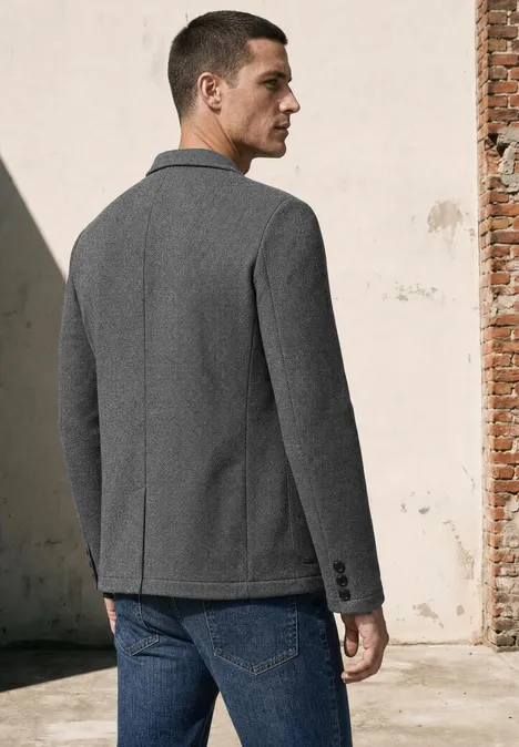 Een man staand van achteren met een grijze blazer en blauwe jeans voor een lichte muur.