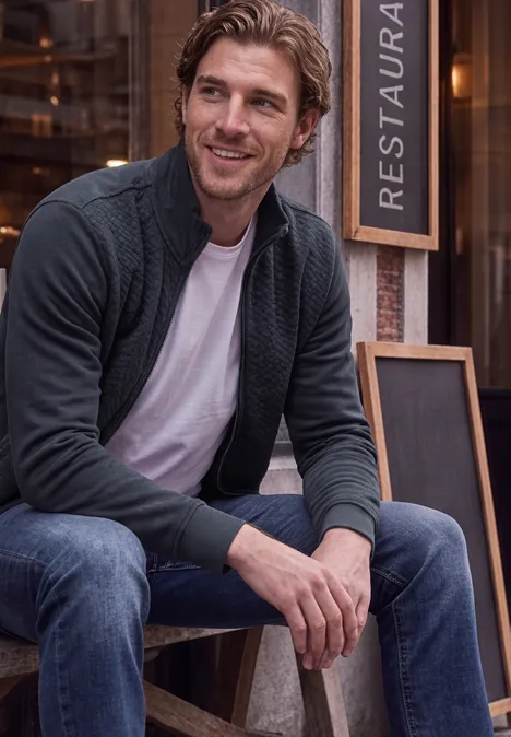 Ein lächelnder Mann sitzt vor einem Restaurant, trägt Jeans, T-Shirt und Strickjacke.