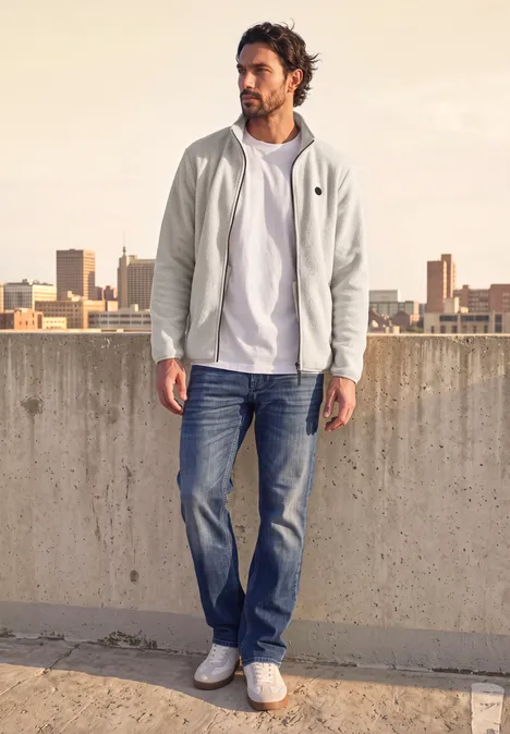 Ein Mann in weißem Hoodie, T-Shirt, Jeans und Sneakern steht auf einem Betonparkdeck mit Stadtblick.
