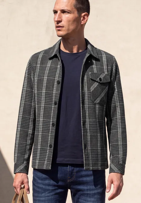 Homme en veste chemise à carreaux gris et noir, t-shirt bleu marine et jean, sur fond clair.