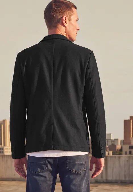 Ein Mann steht mit dem Rücken zur Kamera, trägt einen schwarzen Blazer und schaut auf die Stadtkulisse.