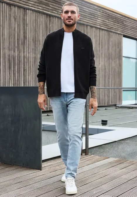 Een bebaarde man in een zwart jasje, wit t-shirt en spijkerbroek staat op een houten terras voor een modern gebouw.