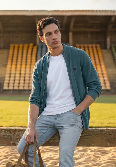 Een jonge man in spijkerbroek en jasje staat met tas voor lege tribunes op een sportveld.