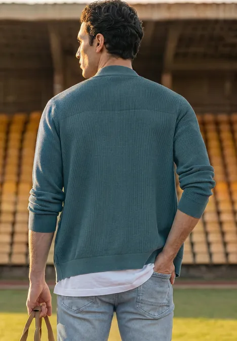 Een man met zijn rug naar de camera staat in een stadion, draagt jeans, trui en houdt een bal vast.