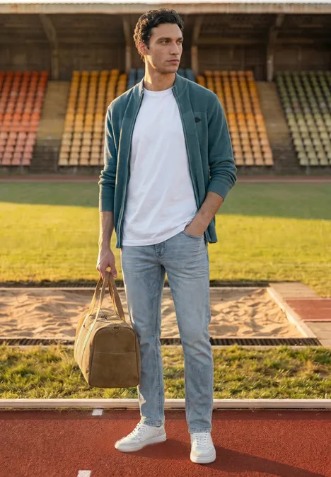 Jonge man in casual kleding met reistas in de hand, op een atletiekbaan met lege tribunes.