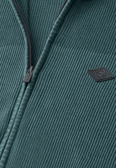 Close-up van een donkergroene, geribbelde gebreide jas met rits en klein logo op de borst.
