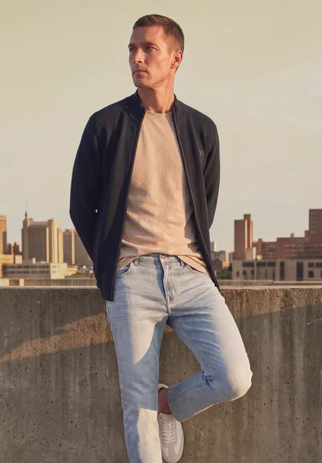 Strickjacke mit Stehkragen und Struktur dark basic blue Ein Mann in Jeans, T-Shirt und Jacke steht lässig auf einem Dach vor einer urbanen Skyline.