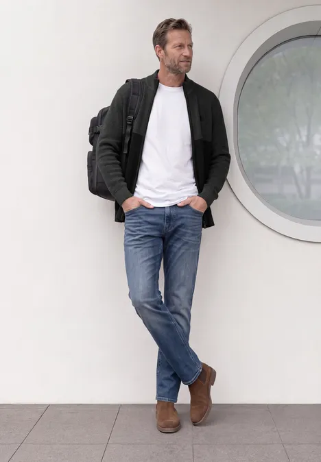 Un homme décontracté appuyé contre un mur près d'une fenêtre ronde, vêtu de jean, tee-shirt blanc, veste et sac à dos.