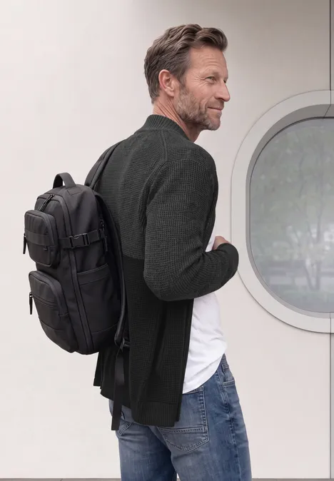 Un homme en jean et blazer porte un sac à dos noir et regarde sur le côté devant un mur blanc avec une fenêtre ronde.