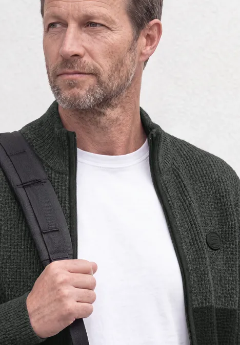 Un homme d'âge moyen à la barbe grise porte un gilet vert, un T-shirt blanc et un sac bandoulière.