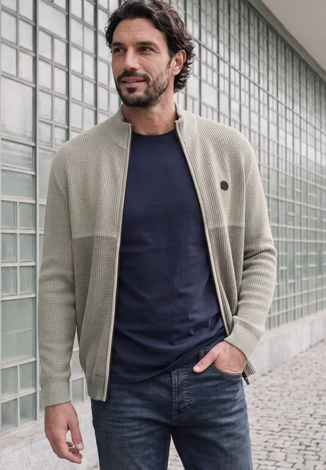 Kardigan z elementami w stylu mouliné i zamkiem błyskawicznym bright beige melange Mężczyzna w beżowym swetrze, granatowym T-shircie i dżinsach stoi przed szklaną ścianą.