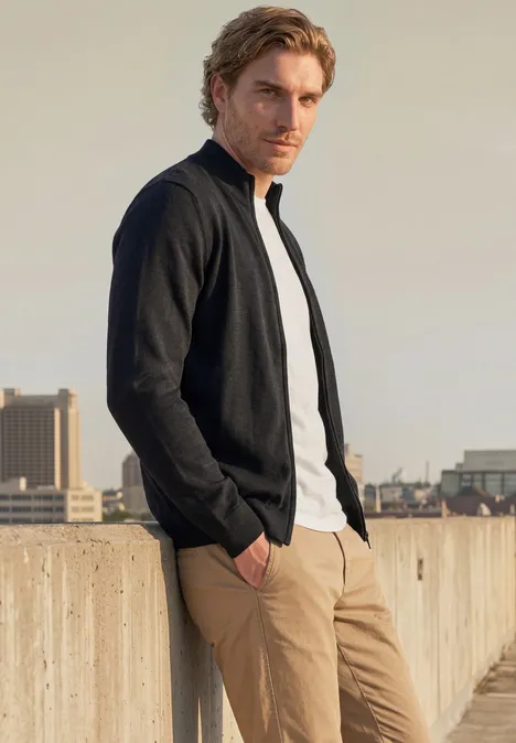 Ein Mann in schwarzer Jacke, weißem T-Shirt und beiger Hose lehnt entspannt an einer Dachbrüstung mit Stadtblick.