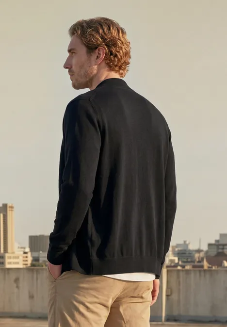 Ein Mann steht auf einem Dach, trägt schwarzen Pullover und beigen Hose, Blick auf die Stadt im Hintergrund.