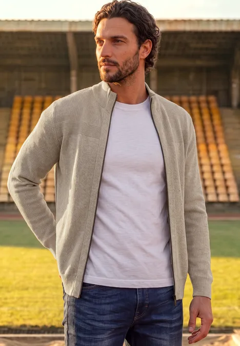 Ein Mann in grauer Jacke, weißem T-Shirt und Jeans steht auf einem Sportplatz vor leeren Tribünen.