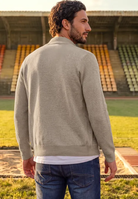 Mann im beigen Pullover und Jeans steht mit dem Rücken zum Betrachter auf einer Laufbahn vor leeren Stadionrängen