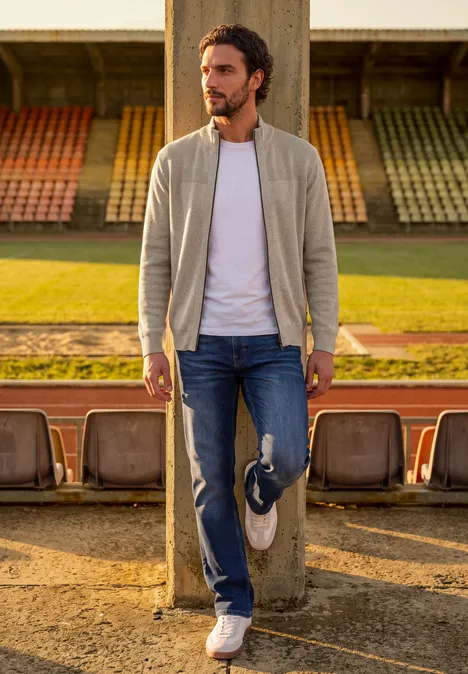 Ein Mann lehnt lässig an einer Säule auf der Tribüne, trägt Bomberjacke, Jeans und hält Turnschuhe.