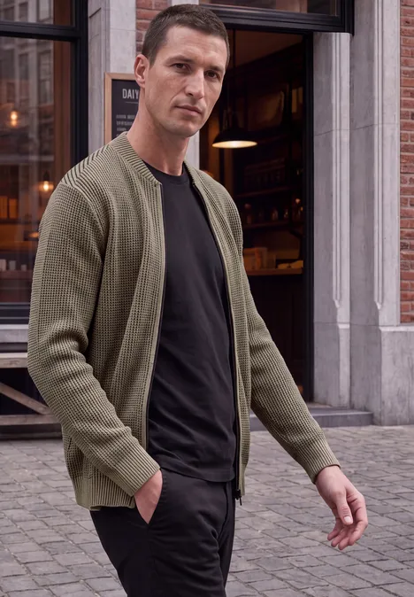 Ein Mann in grüner Strickjacke und schwarzem T-Shirt läuft vor einem Café in der Stadt.