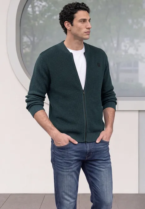 Ein Mann trägt eine dunkelgrüne Strickjacke mit Reißverschluss, weißes T-Shirt und Jeans vor einem runden Fenster.