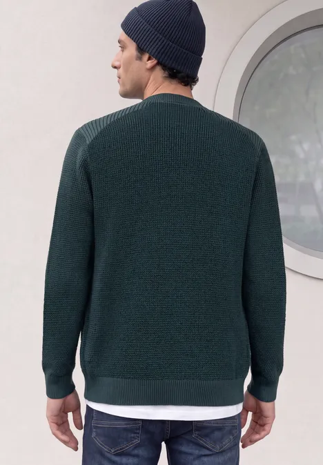 Ein Mann von hinten, trägt dunkelgrünen Pullover, weiße Unterziehschicht und Strickmütze vor rundem Fenster.