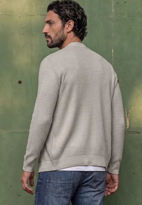 Een man in profiel draagt een beige shirt met wafelstructuur en jeans voor een groene wand.