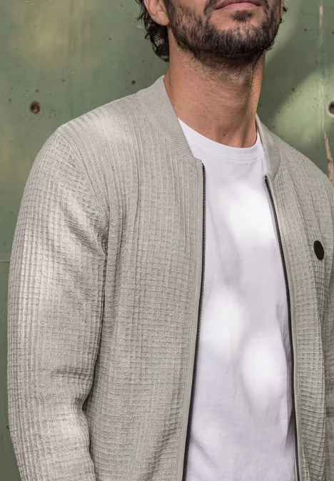 Een man draagt een wit T-shirt onder een lichtgrijze gebreide cardigan voor een groene achtergrond.