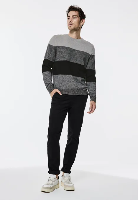 Mann trägt gestreiften Pullover, schwarze Hose und weiße Sneaker vor weißem Hintergrund.