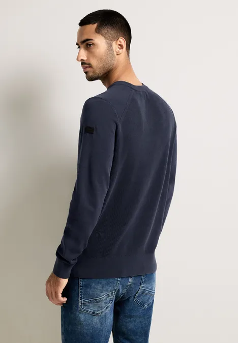 Mann in dunkelblauem Pullover und Jeans steht seitlich zur Kamera.