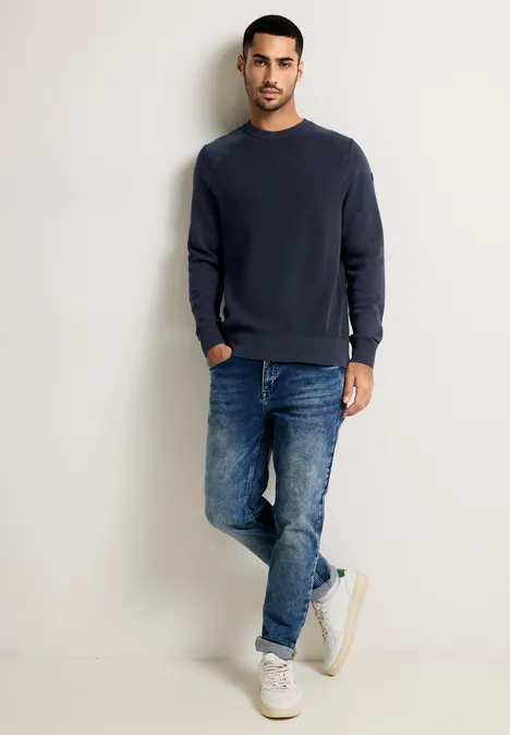 Mann in dunkelblauem Pullover, Jeans und weißen Sneakers steht vor heller Wand.