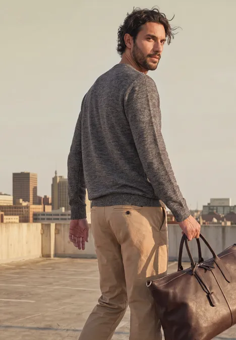 Ein Mann auf einem Parkdeck trägt grauen Pullover, bequeme Hose und hält eine braune Reisetasche.