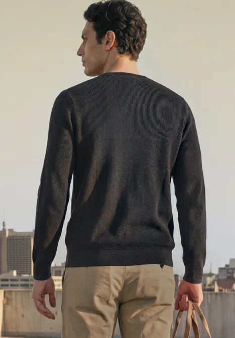 Ein Mann steht mit dem Rücken zur Kamera auf einem Dach, trägt dunklen Pullover, helle Hose und hält eine Tasche.